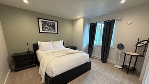 A bedroom with a bed, nightstands, a bedside table, a fan, and a window with curtains. - Cozy Two-Storey Suite in Maple Bay (Duncan)