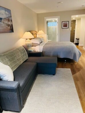 A hotel room with a sofa, a bed, a nightstand with lamps, a painting on the wall, and a door leading to another room. - Oceanside Garden Studio B: Pool & Hot tub (Nanoose Bay)
