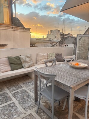 Eine Dachterrasse mit Tisch und Stühlen, einer gepolsterten Bank und Blick auf die Skyline im Sonnenuntergang.