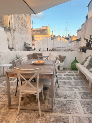 Eine Terrasse im Freien mit einem Holztisch und Stühlen, einem grünen Blumentopf und Blick auf Gebäude im Hintergrund.