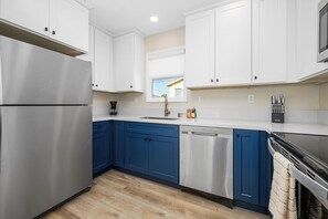 A modern kitchen with white cabinets, blue lower cabinets, stainless steel appliances, and a window with a view of a house. - Beachfront Studio Steps to Main Street (Seal Beach)