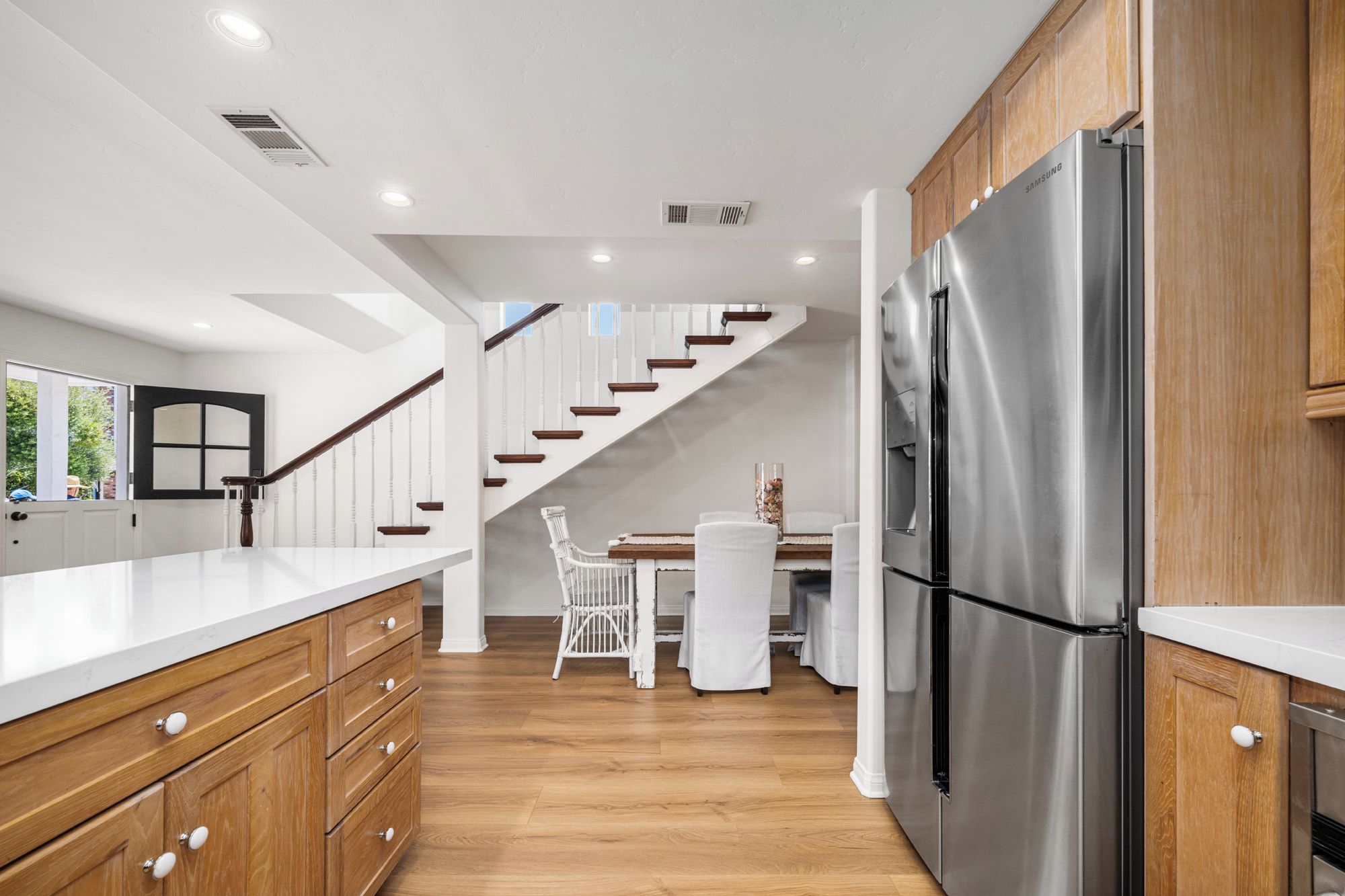 A modern kitchen with a stainless steel refrigerator, wooden cabinets, a white countertop, and a staircase leading to a dining area.