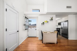 A modern kitchen with white cabinetry, a stainless steel refrigerator, and a dining area with a wooden table. - Coastal Living on Balboa Island (Newport Beach)