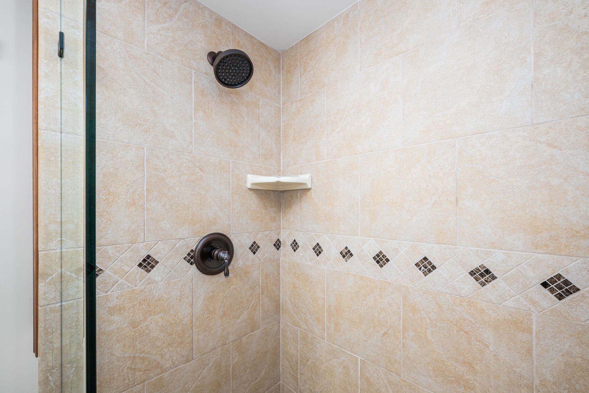 Área de chuveiro com azulejos bege, chuveiro manual, chuveiro fixo na parede e uma pequena prateleira.