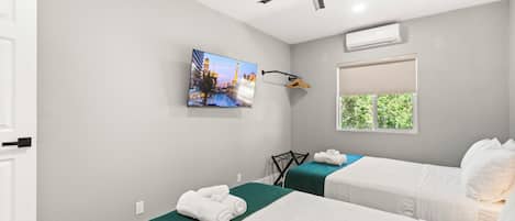 A hotel room with two beds, a ceiling fan, an air conditioning unit, a TV on the wall, and a window with a view of greenery.