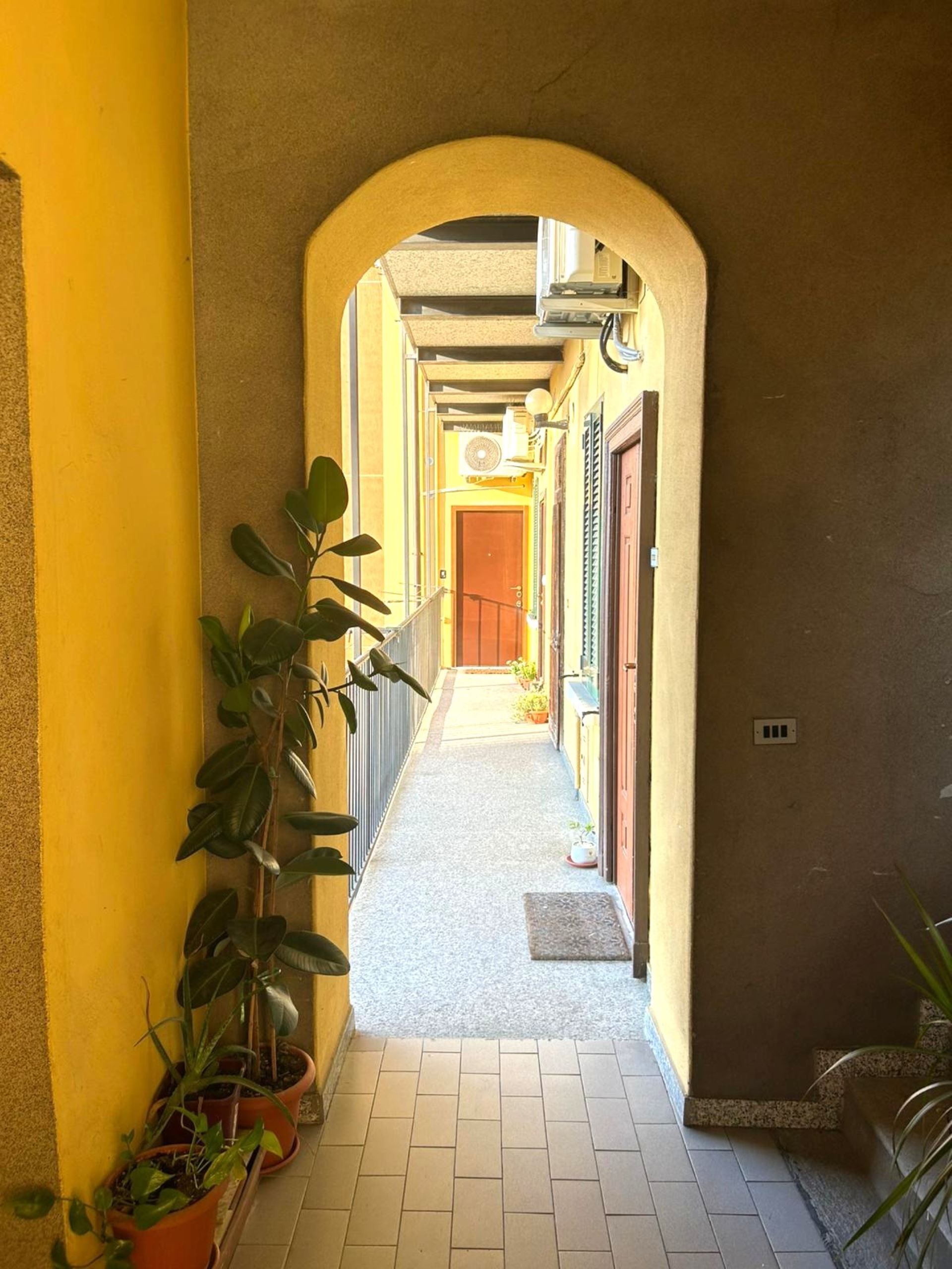 A narrow hallway with a tiled floor, potted plants, and a door at the end.