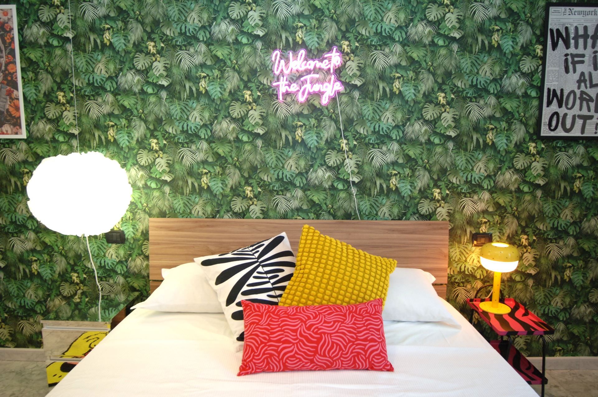 A bed with a wooden headboard, multiple pillows, a neon sign, a lamp, and a wall with a leafy pattern.