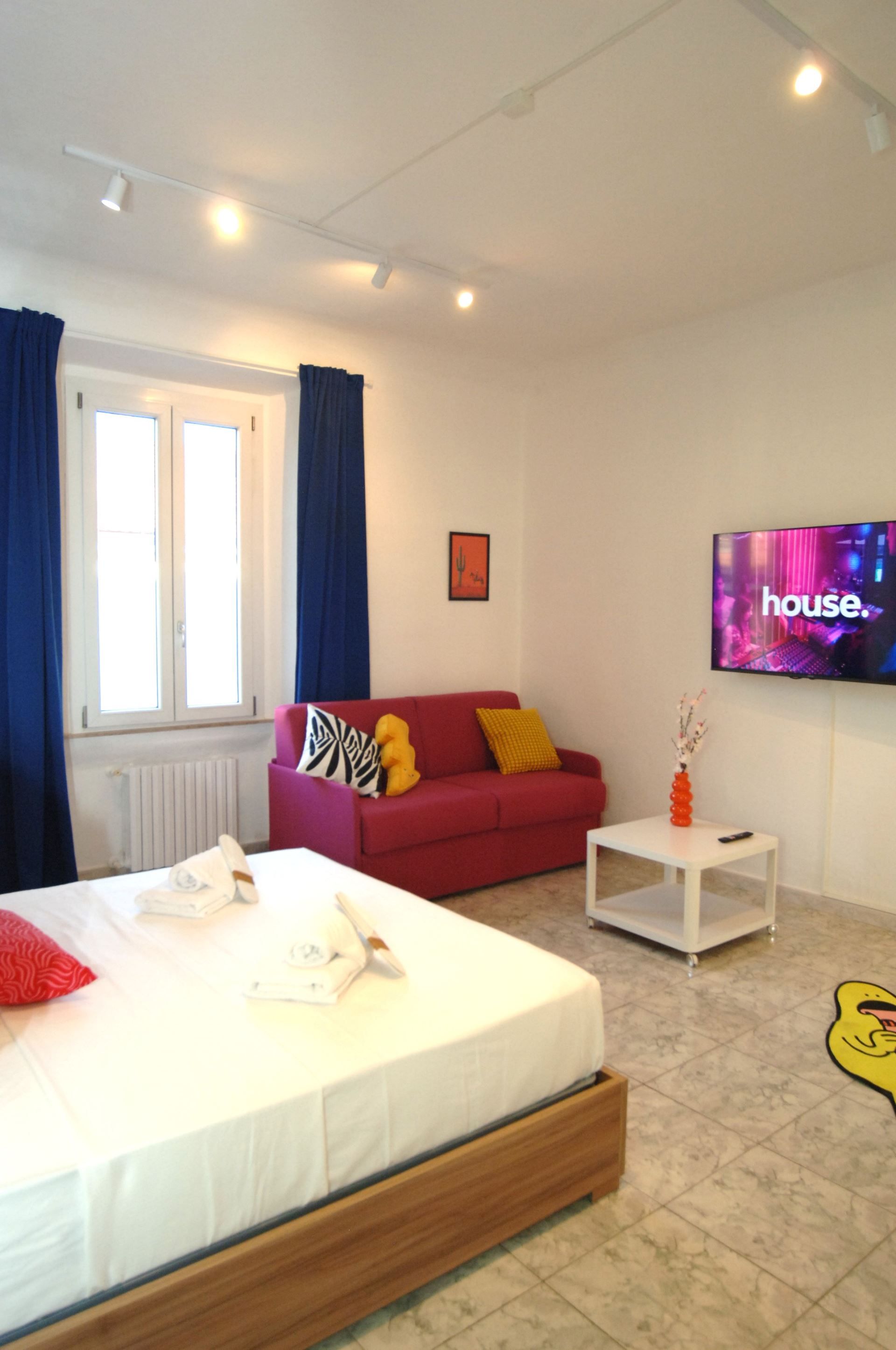 A bedroom with a bed, a red sofa, a television, and a window with curtains.