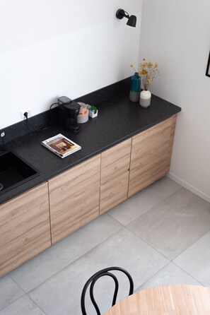 Una cocina moderna con encimera negra, armarios de madera, cafetera, una revista y un florero con flores.