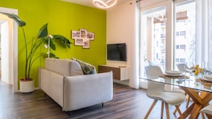 Ein modernes Wohnzimmer mit einer Couch, einem Esstisch mit Stühlen, einem Fernseher und einem großen Fenster mit Blick auf Gebäude.