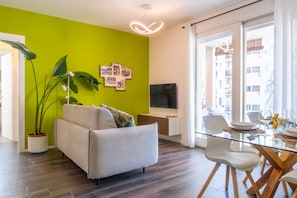 A modern living room with a sofa, a dining table with chairs, a TV, and a large window with a view of buildings. - Green Apartment by MARTINI's Home (Milano)