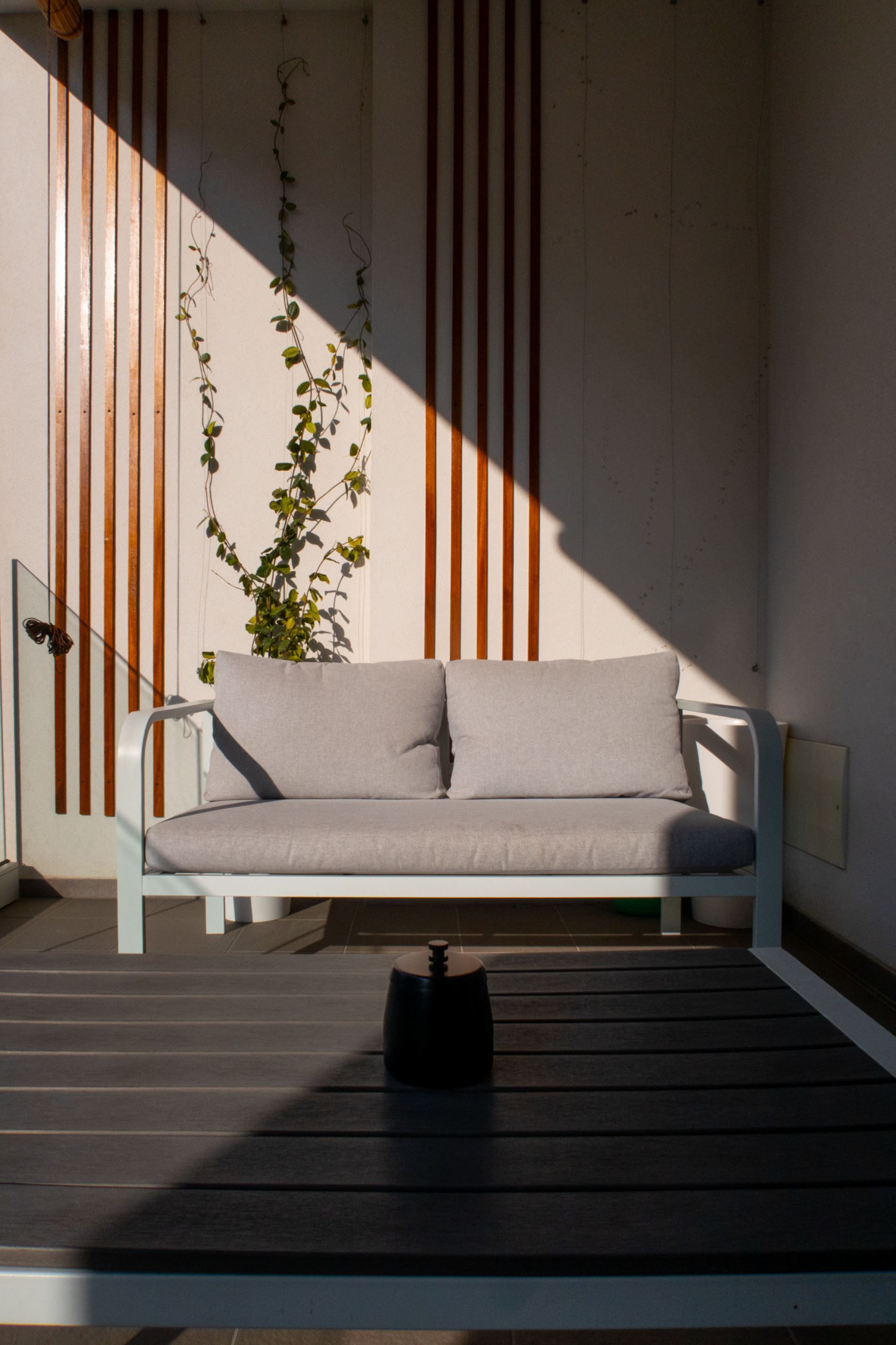 A modern outdoor seating area with a white sofa and a small table, set against a wall with vertical wooden slats and a climbing plant.