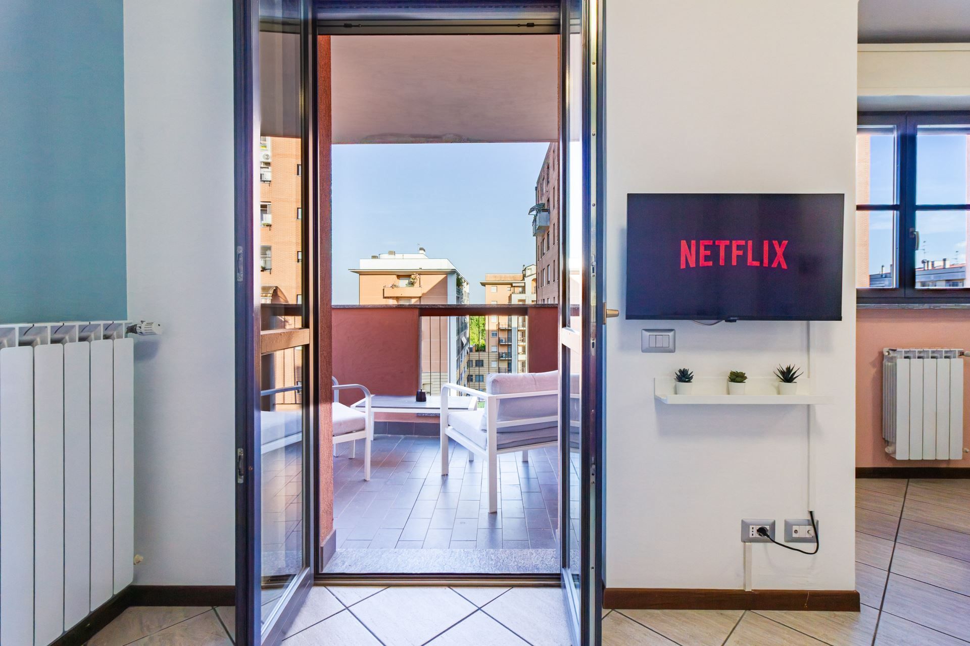 A hotel room with a sliding glass door leading to a balcony, a television on the wall, and a view of the cityscape.