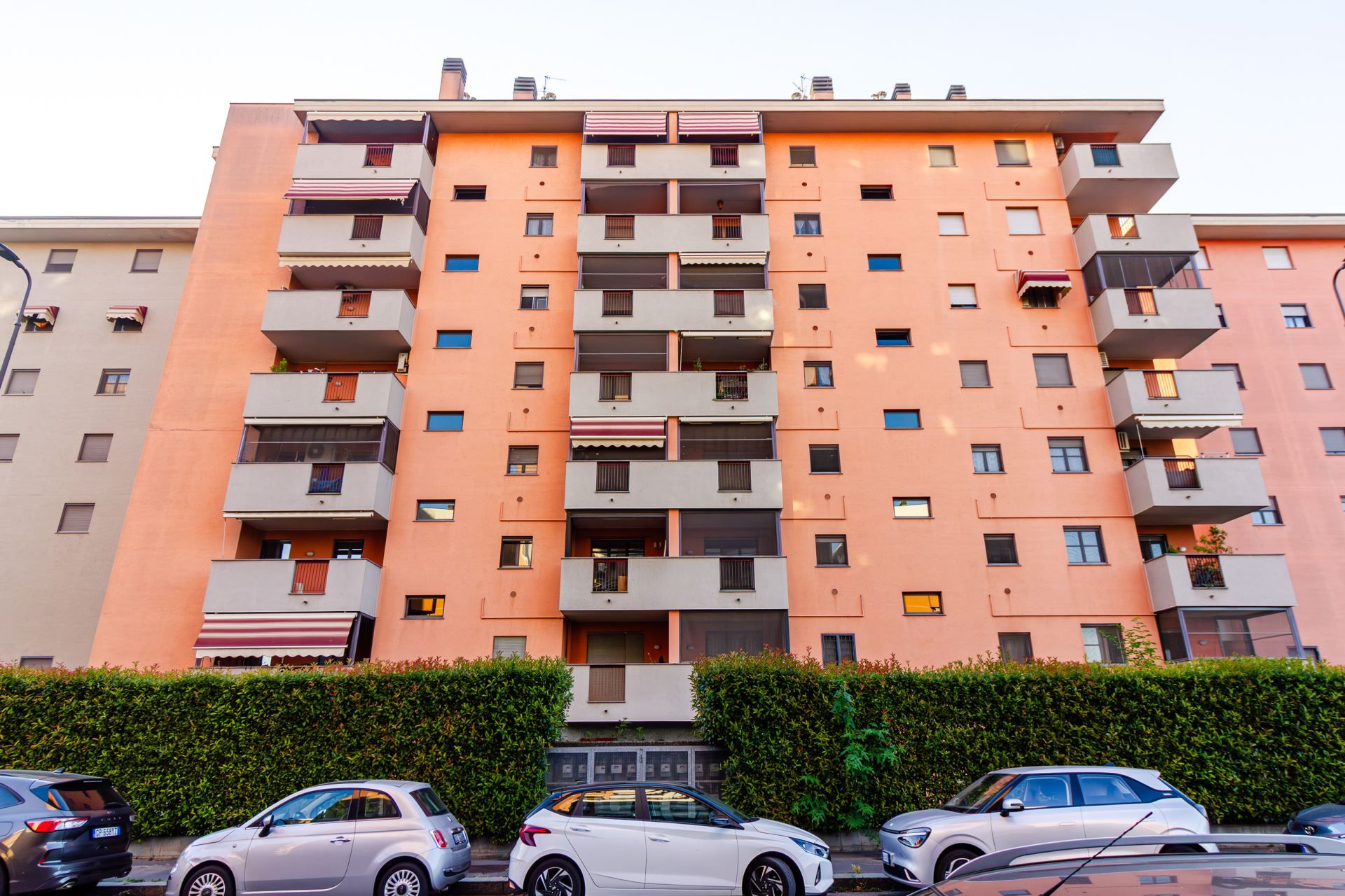 Un edificio residenziale a più piani con balconi, un'area di parcheggio e un siepe antistante.