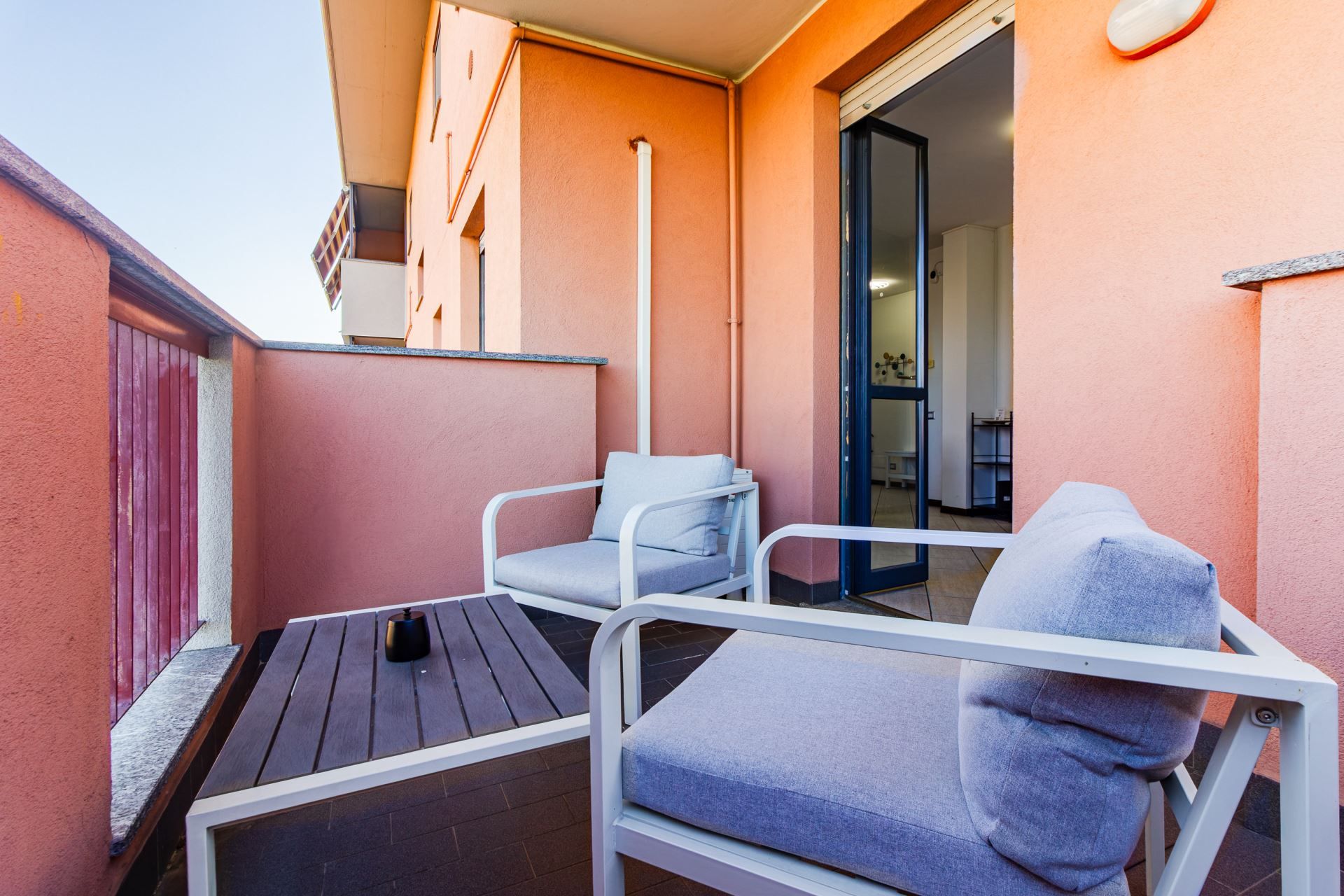 Een balkon met twee witte metalen stoelen, een houten tafel, een rode deur en een witte muur.