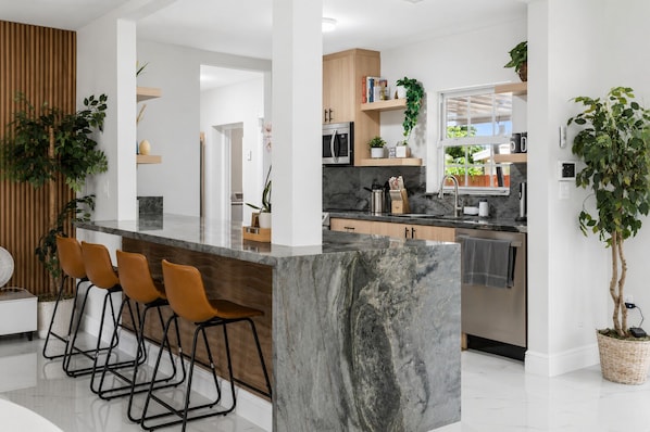 A modern kitchen with a marble island, wooden chairs, and potted plants. - Luxury 5BR Heated Pool, Game Room, Sandy Beach Area & Basketball (Miami)