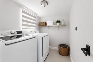 A laundry room with a front-loading washer and dryer, a wicker basket, and a shelf with towels and a plant. - Luxury 5BR Heated Pool, Game Room, Sandy Beach Area & Basketball (Miami)