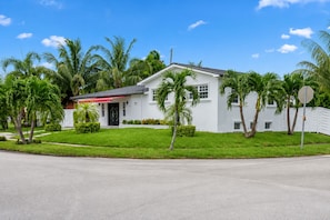 A single-story white house with a red awning, surrounded by well-maintained grass and palm trees. - Luxury 5BR Heated Pool, Game Room, Sandy Beach Area & Basketball (Miami)
