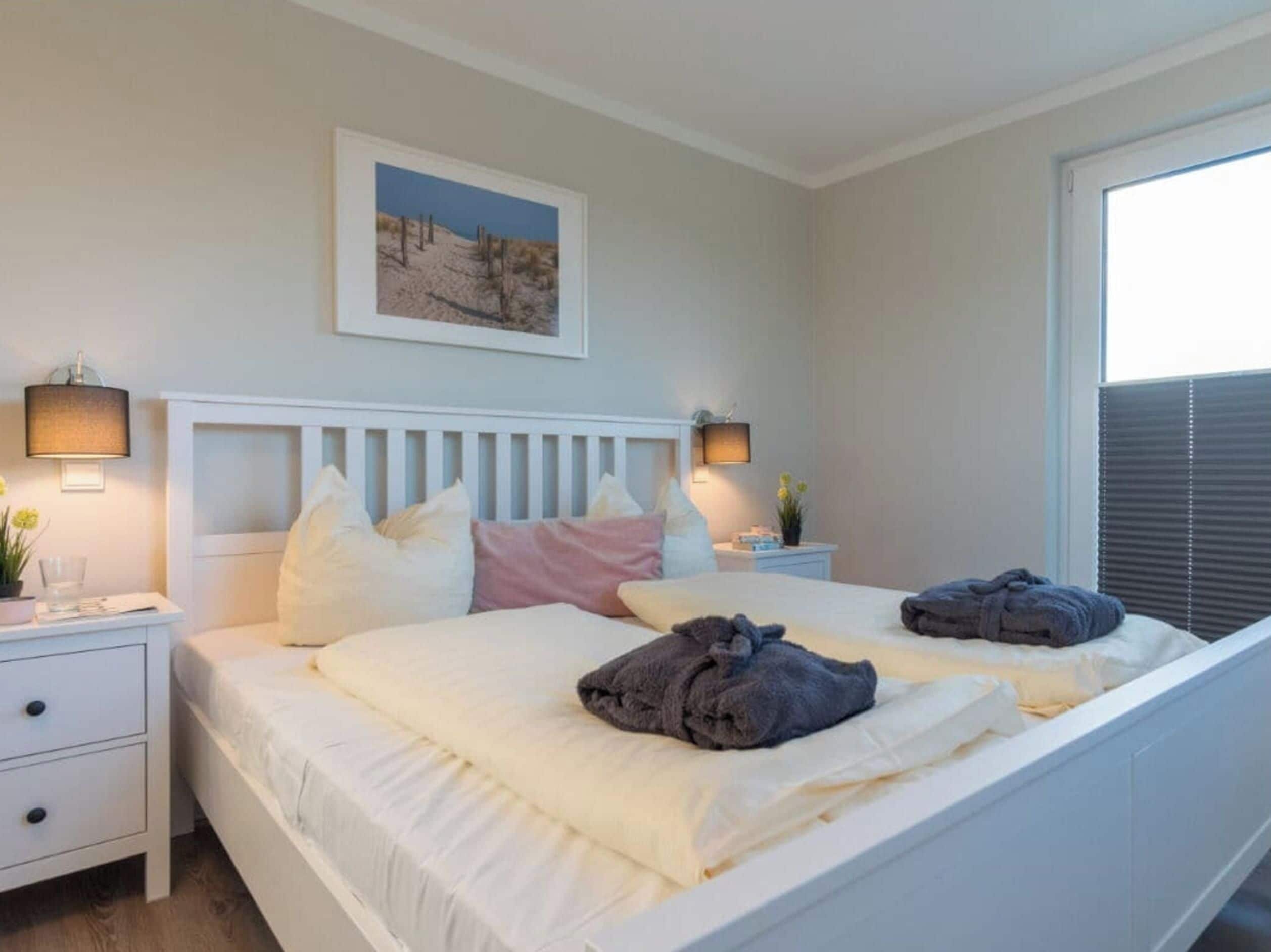 Une chambre à coucher avec un lit blanc, deux tables de chevet avec des lampes, un tableau encadré au mur et une fenêtre avec des stores.