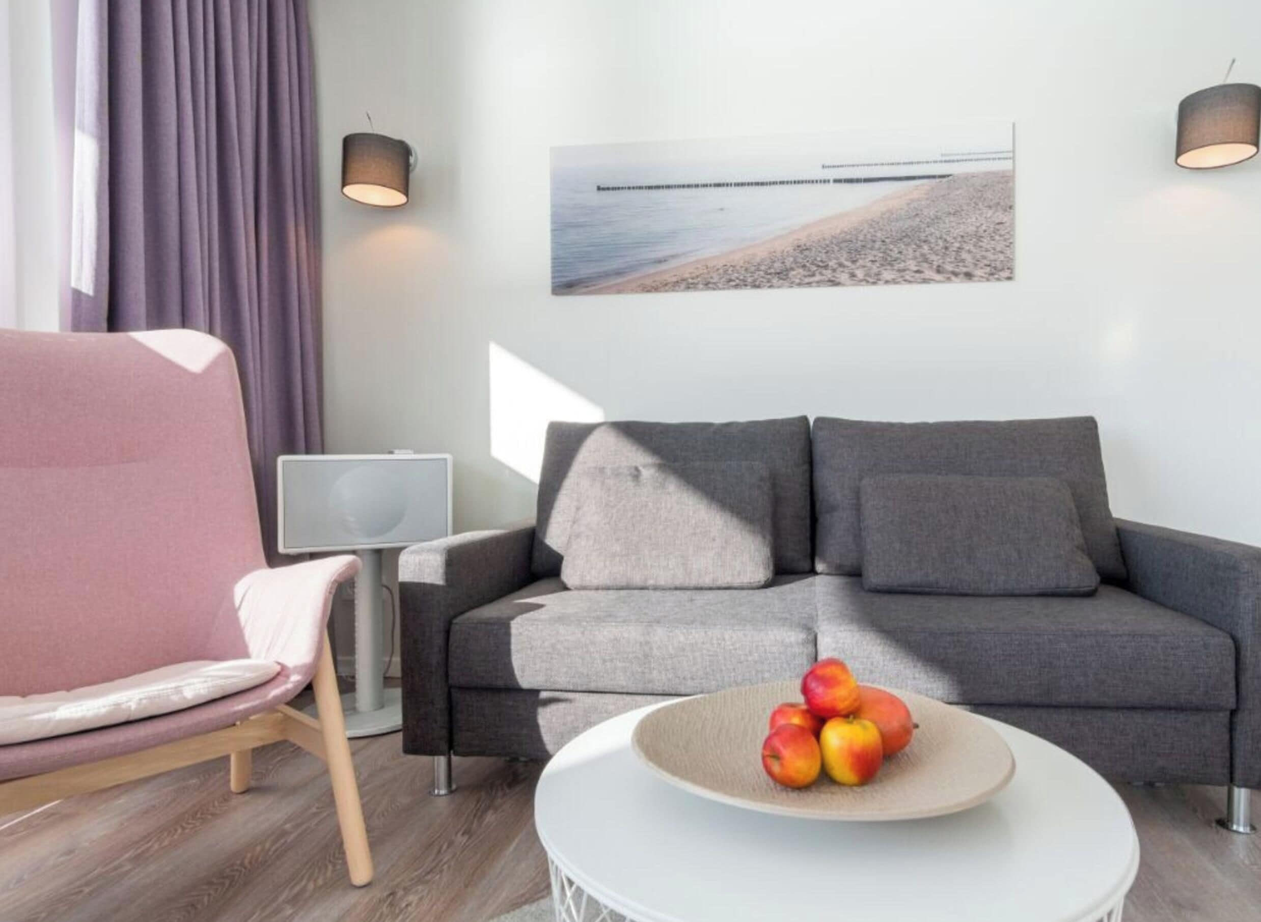 Una sala de estar moderna con un sofá gris, un sillón rosa, una mesa redonda blanca con fruta y una lámpara de pared.