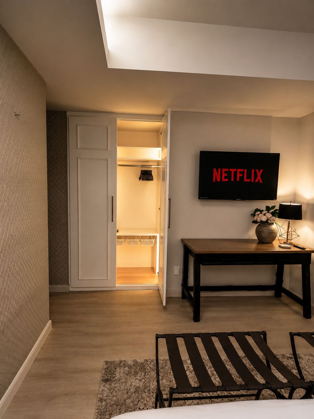 Ein Hotelzimmer mit einem Bett, einem Schreibtisch mit Lampe, einem Fernseher, der 'NETFLIX' anzeigt, und einem Kleiderschrank mit Kleidung.
