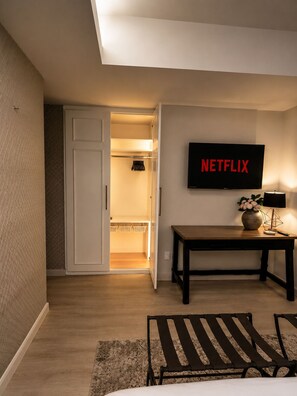 Ein Hotelzimmer mit einem Bett, einem Schreibtisch mit Lampe, einem Fernseher, der 'NETFLIX' anzeigt, und einem Kleiderschrank mit Kleidung.