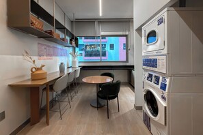 Eine moderne Küche mit integriertem Waschmaschine, eine Essecke mit rundem Tisch und Stühlen sowie Blick auf ein farbenfrohes Gebäude durch ein Fenster.