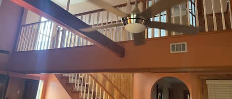 Un interior de varios niveles con una escalera, vigas de madera y un ventilador de techo.
