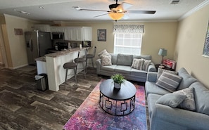 A compact living space with a kitchenette, a grey sofa, a coffee table with a plant, and a colorful rug.