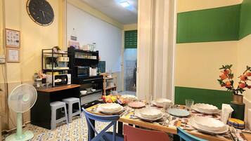 A dining area with a table set for four, chairs, a fan, a clock, and a kitchen area with appliances and a shelf.