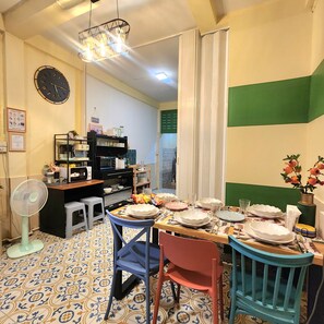 Un coin repas avec une table dressée pour quatre personnes, des chaises, un ventilateur, une horloge et un espace cuisine équipé d’appareils électroménagers et d’une étagère.