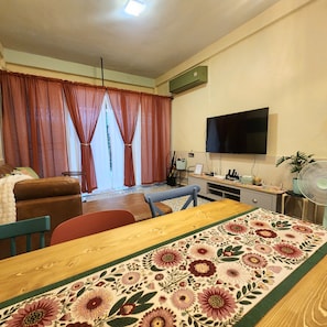 Ein Wohnzimmer mit einer Couch, einem Esstisch mit Blumenläufer, einem an der Wand befestigten Fernseher und einem Ventilator auf einem Beistelltisch.