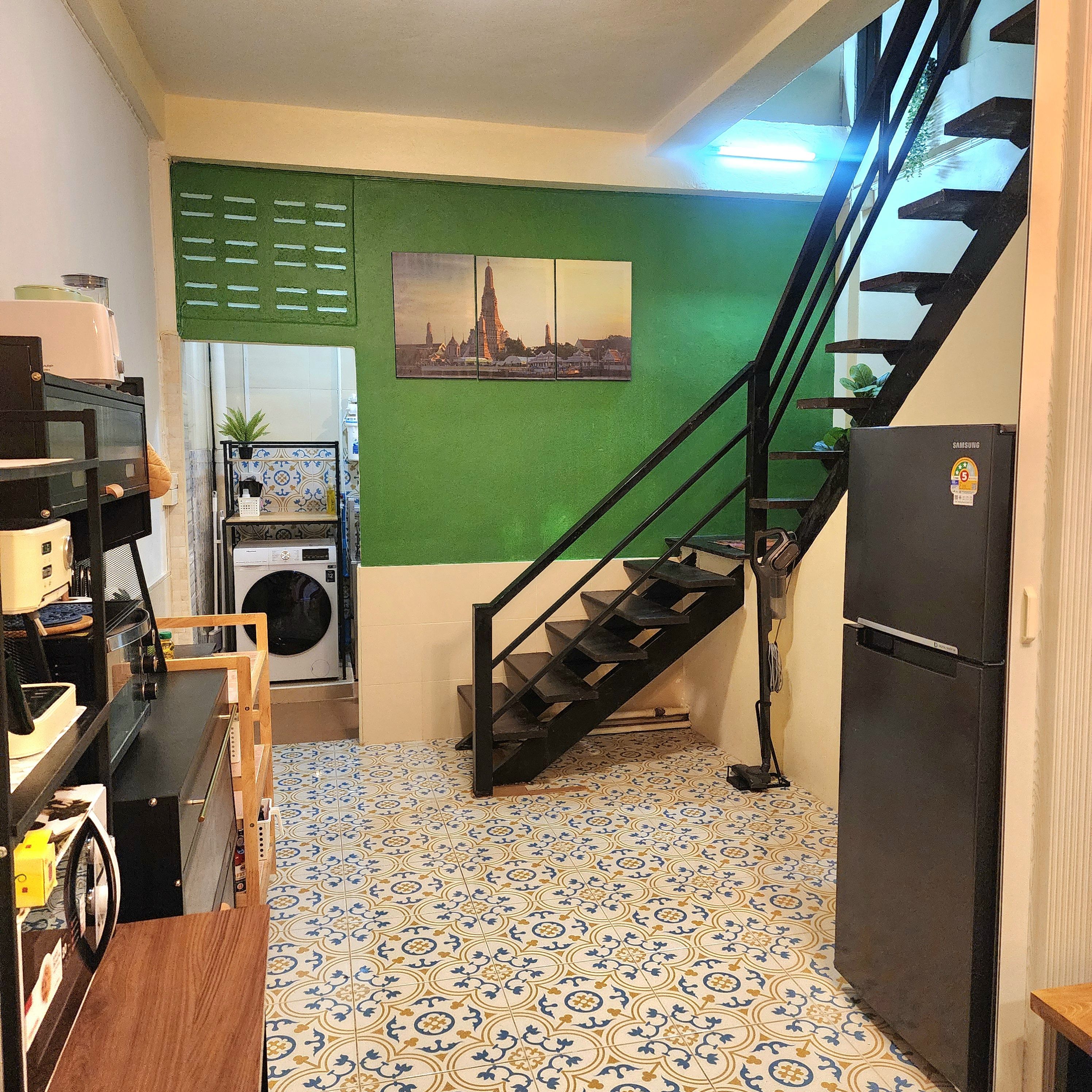 A kitchen with a washing machine, a Samsung refrigerator, a staircase, and a green wall with a cityscape picture.