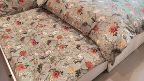 Ein Bett mit floralem Bettzeug und passendem Kopfkissen, ein Nachttisch mit Lampe sowie ein Fenster mit Vorhängen.