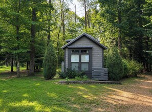 A small cabin with a sloped roof, surrounded by dense trees and a well-maintained lawn.