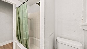 Un baño con inodoro blanco, ducha con cortina de cuadros verdes y pared de azulejos.