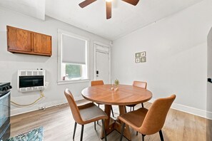 En matplats med ett runt träbord och fyra stolar, en takfläkt, ett fönster med persienner och en väggmonterad ugn.