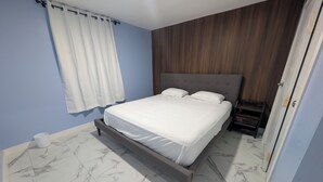 Quarto com cama, mesa de cabeceira, cabeceira de madeira, cortina branca e piso de mármore.