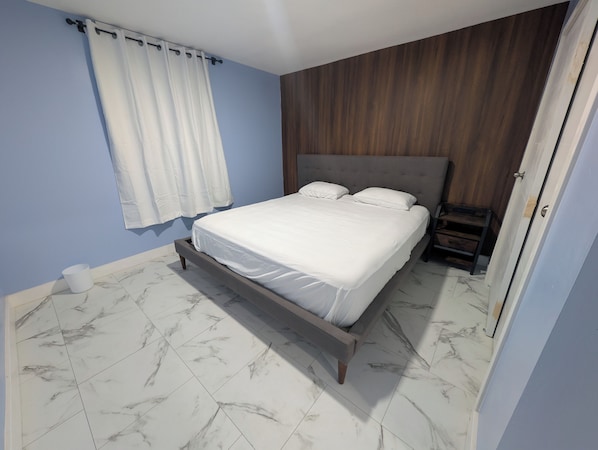 A bedroom with a bed, a nightstand, a wooden headboard, a white curtain, and a marble-tiled floor.