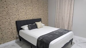 Uma cama com cabeceira escura, roupa de cama branca e manta cinza.