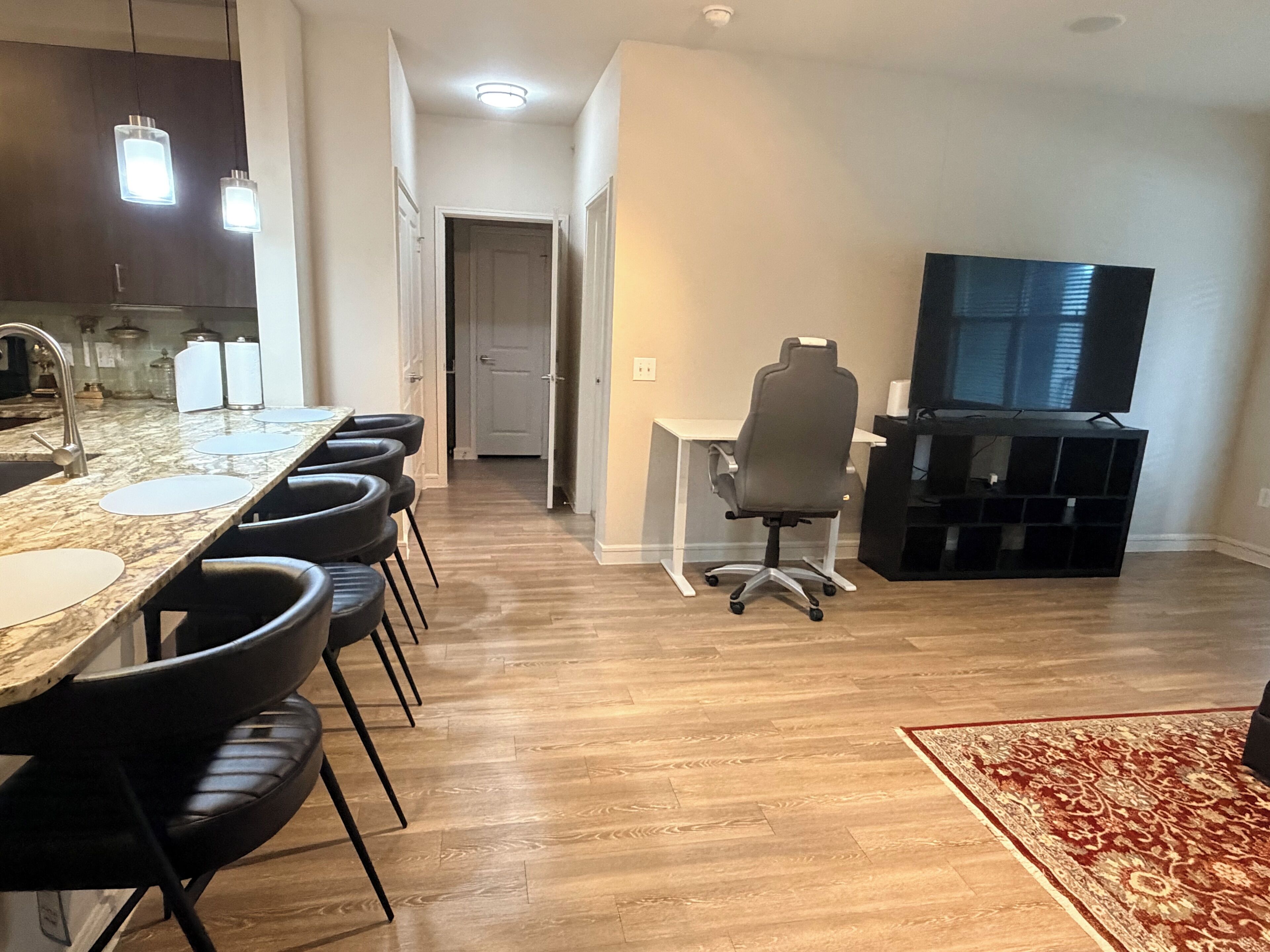 A modern kitchen with a marble countertop, a dining area with chairs, a TV, and a carpeted floor.