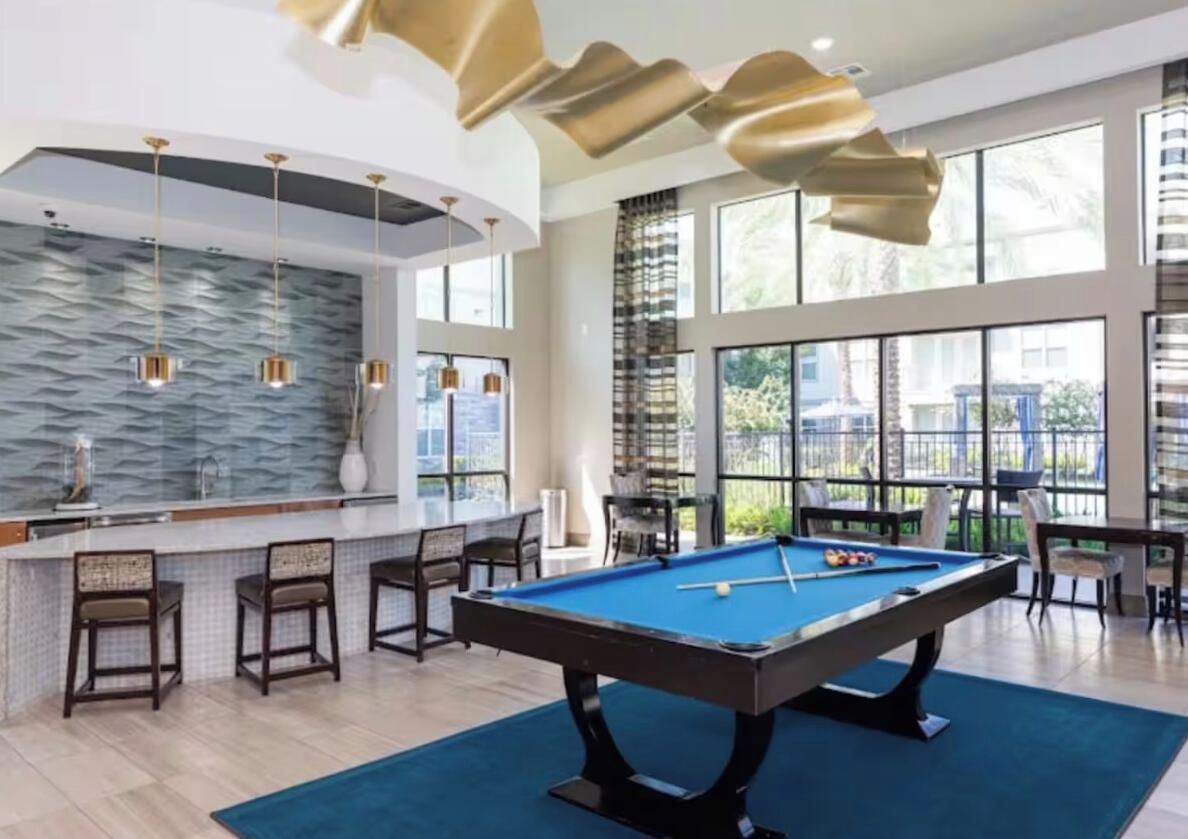 A pool table in a well-lit room with large windows, a bar area with stools, and a modern ceiling design.