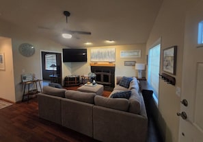 A living room with a sectional sofa, a fireplace, a flat-screen TV mounted on the wall, and a ceiling fan.