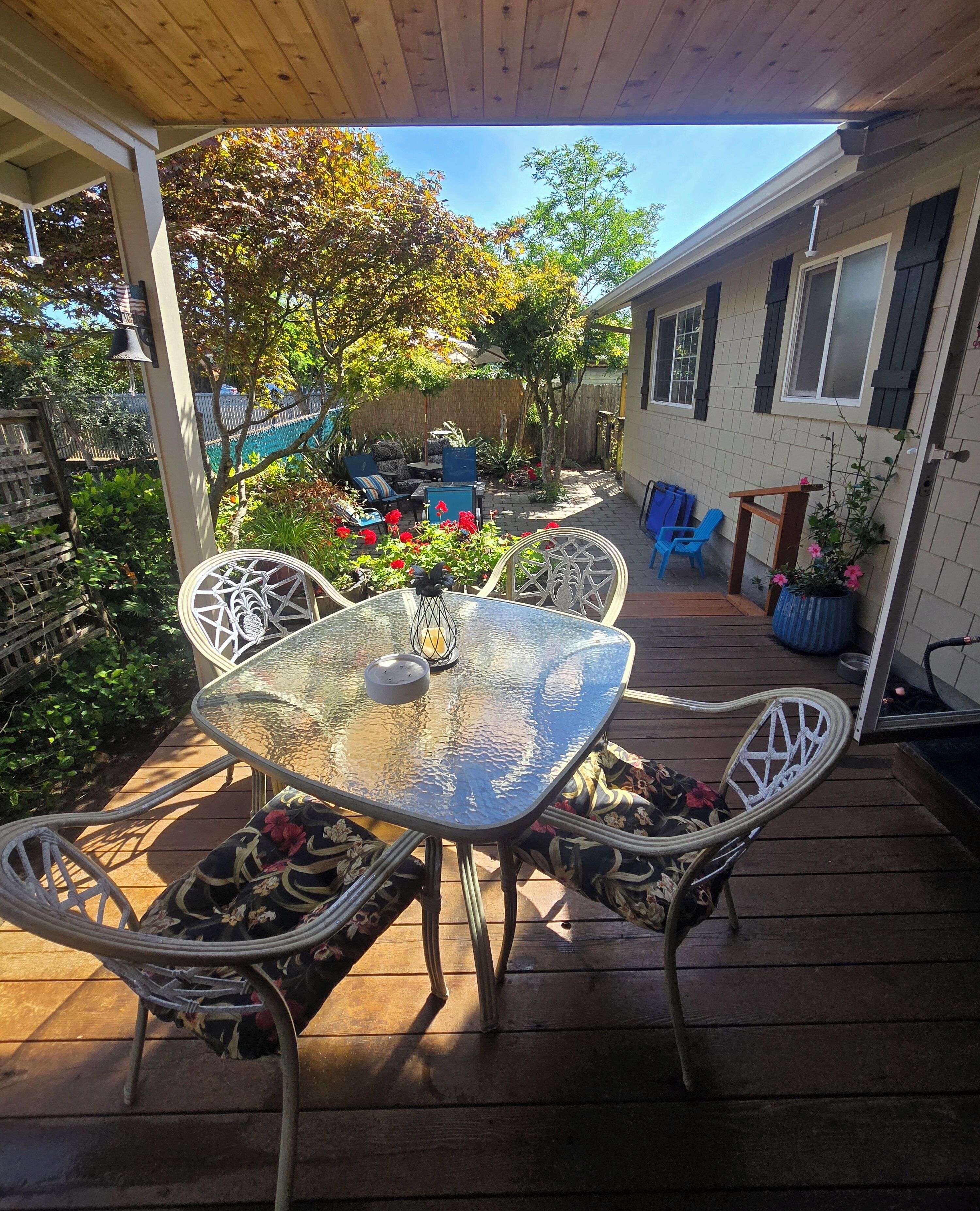 An outdoor patio with a glass table and metal chairs, surrounded by a wooden deck and a garden with various plants and flowers.
