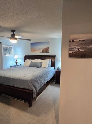 A bedroom with a bed, a ceiling fan, a nightstand with a lamp, and two wall-mounted paintings.