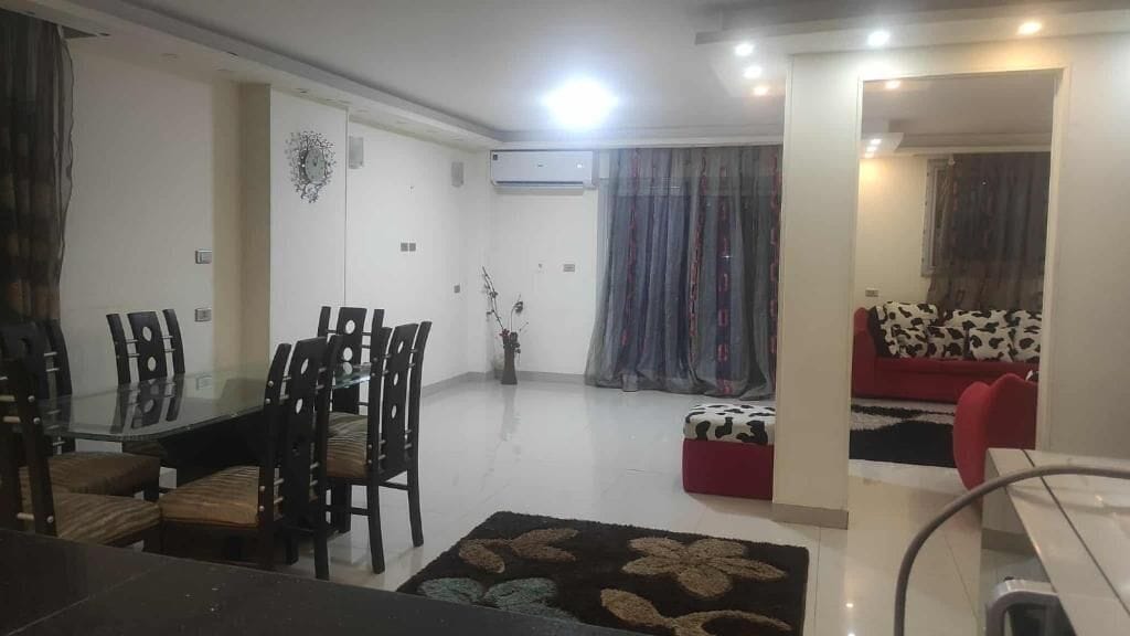 A living room with a glass dining table, chairs, a sofa, and a red ottoman.