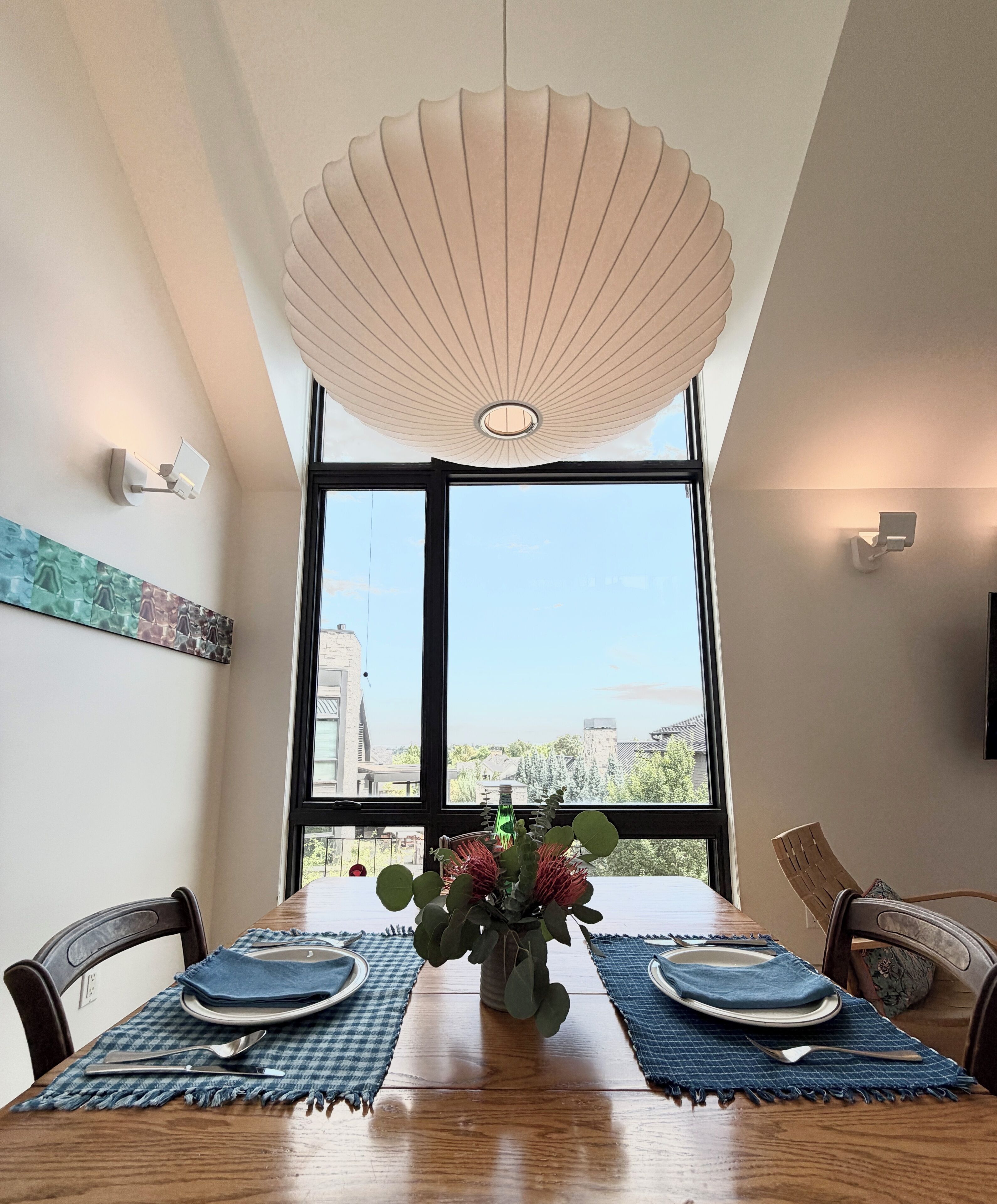 Uma sala de jantar com mesa posta para dois, luminária decorativa no teto e vista através de grandes janelas.