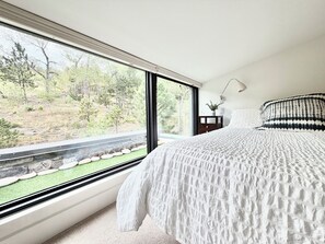 A bedroom with a large window, a bed with a white textured bedspread, a bedside table with a lamp, and a view of a forested area. - Walk to Sundance Film Festival+ Downtown Boulder- Private Guest House (Boulder)