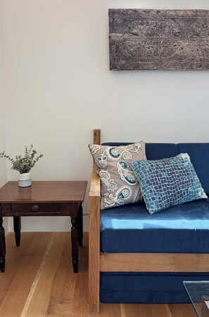 Ein Zimmer mit einem Holztisch, einem blauen Sofa mit gemusterten Kissen und einem Wandbild.