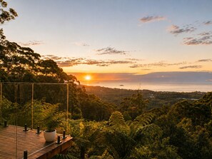 Eine Holzterrasse mit Blumentöpfen, mit Blick auf eine üppige Landschaft und einen Sonnenuntergang über dem Wasser.